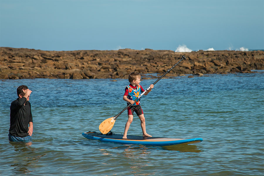 A child is standing on a SUP board, holding a paddle. An adult is nearby, watching. 