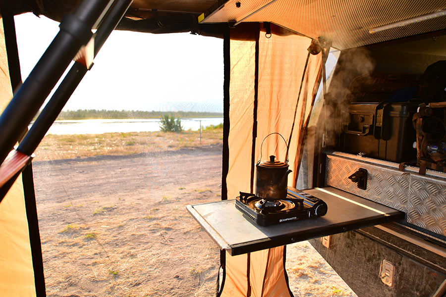 A kettle on a portable camp stove beneath a 4WD awning, letting out steam as the sun rises. 