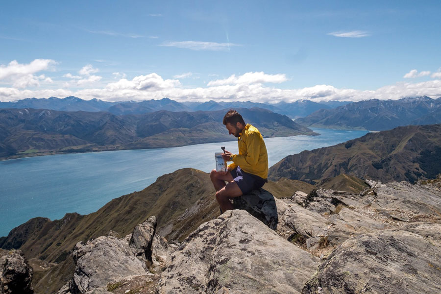 A man sitting on a mountain peak, eating a packet of Radix. 