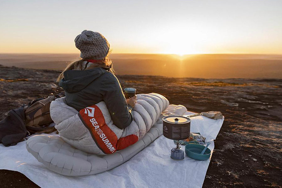 A woman in a beanie sits rugged up on a sleeping mat holding a mug, with a hiking stove to her right, looking out to the sunrise. 