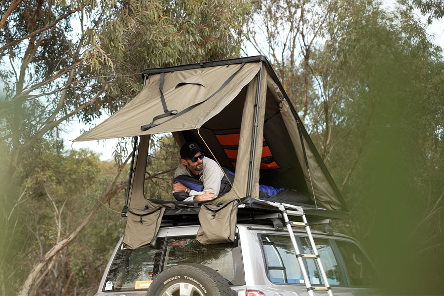 Combining slender builds with outdoor durability for truly elevated camping experiences! Ben from Snowys in a sleeping bag, looking out from his Inspired Overland Roof Top Tent in the bush.
