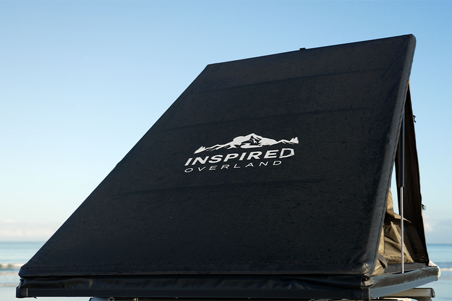 The Lightweight Rooftop Tent range by Inspired Overland elevates every outdoor adventure! Inspired Overland Roof Top Tent, with branding facing the camera and the beach in the backdrop.