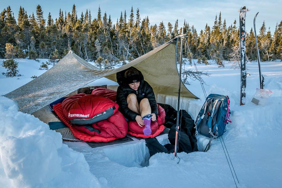 A woman pulling on her socks in the snow. She is sitting on a red sleeping bag under a tarp, and her skies are sticking out of the snow to her left. 