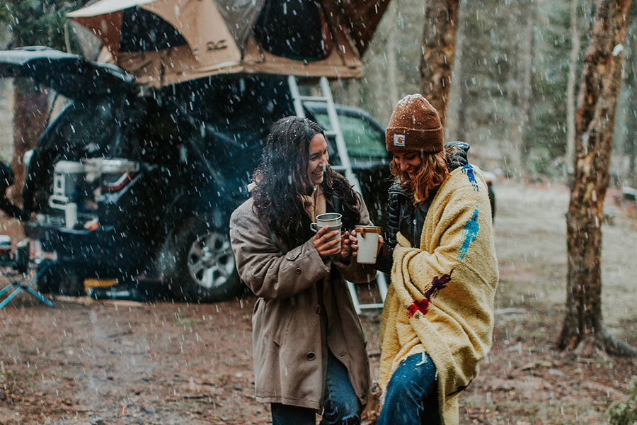 Two women are rugged up, holding coffee cups as light snow falls. 