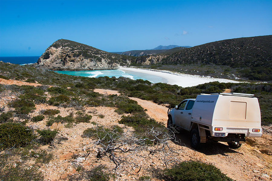 A 4WD is parked on a red rubbly track overlooking bright blue ocean waters and white sand. 