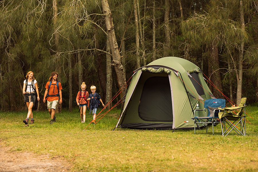 A green tent with two camp chairs and a family of hikers returning to their site.
