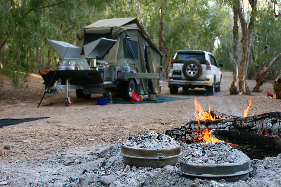 A campfire with coals on top of Trangias, with a 4WD camp setup in the background.