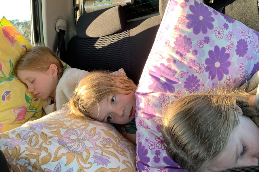 Three kids asleep in the back seat of the car with pillows. 