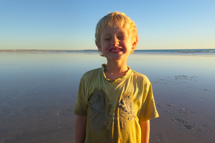 A young boy smiling, covered in wet sand on a beach at sunset. 