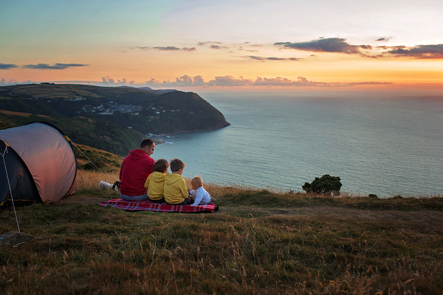 A father and his children are sitting on a picnic rug overlooking the sunset on the ocean, from the top of a cliff. The tent is standing nearby. 