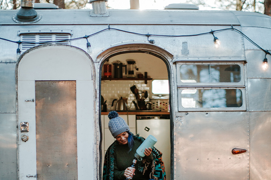 A woman in a beanie sits on a step outside her van, strung with lights.