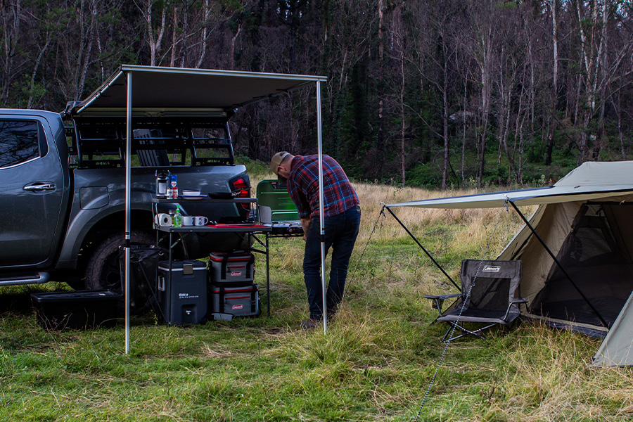 Ben and Lauren decode what's driving the camping cravings! A man is setting up his camp kitchen from the side of his 4WD under an awning, with a tent and chair erect behind him.