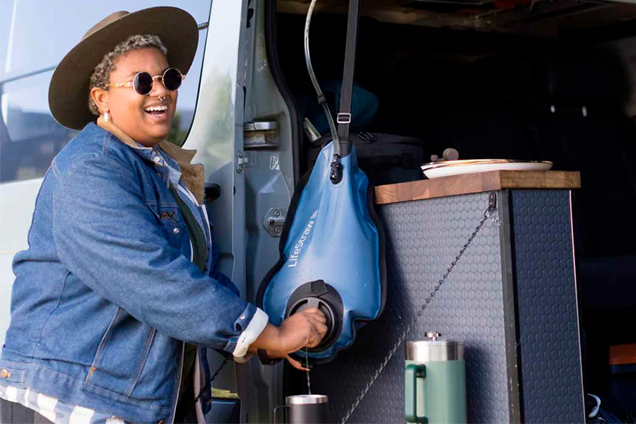 A camper wearing sunglasses, a hat, and a blue jacket smiling while filling their travel cup with water from a LifeStraw pouch. 