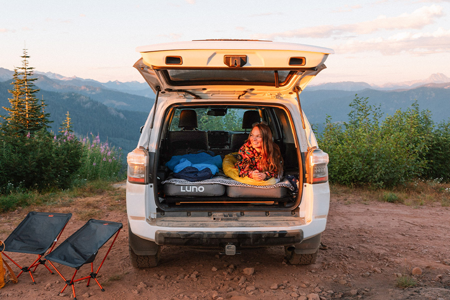 A woman lies on her front in the back of her vehicle with the back lifted, on top of two Luno mattresses, smiling in the sunset on the mountaintop.